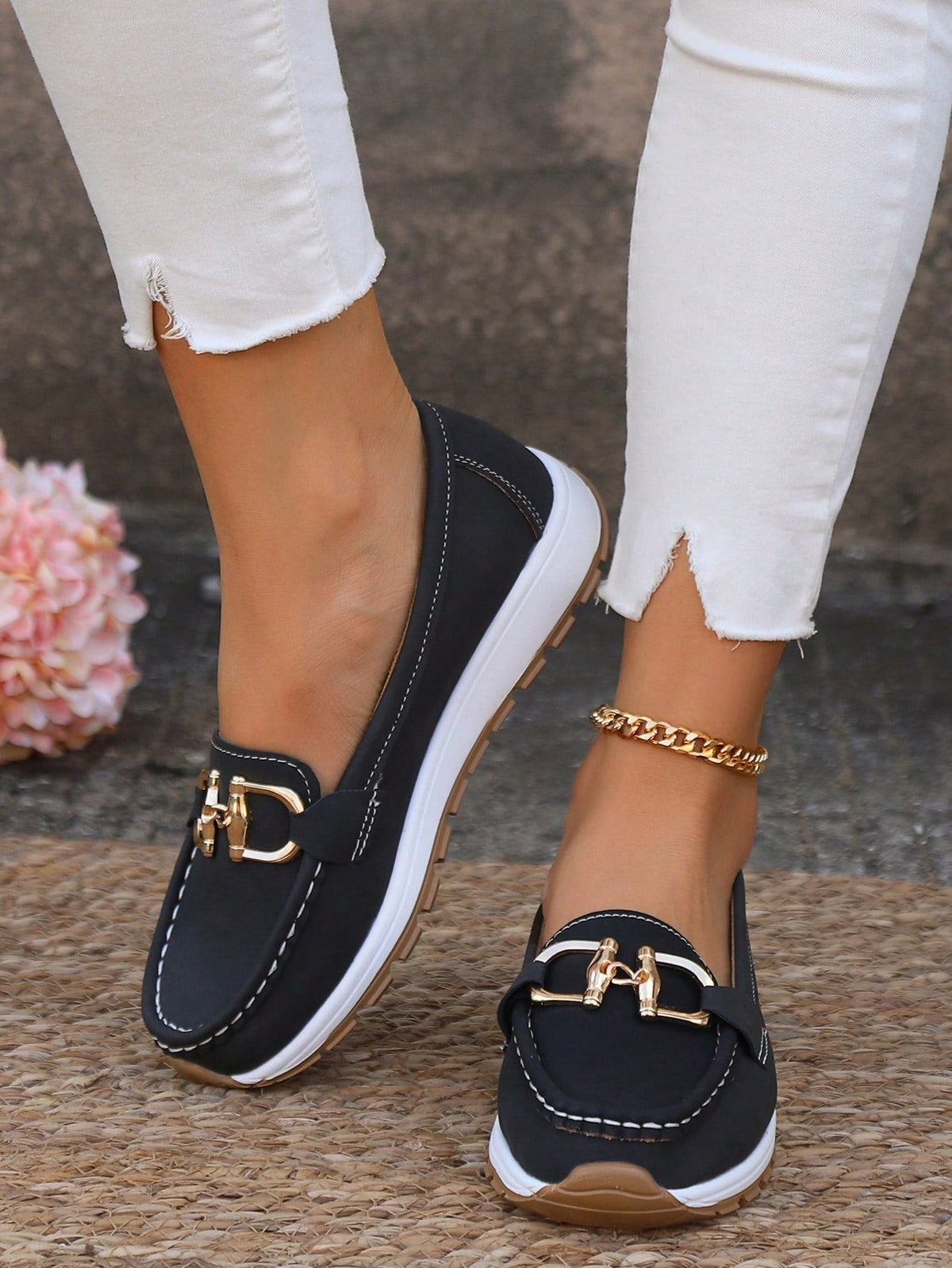 Mocasines negros con plataforma blanca, suela dentada y detalle metálico dorado, combinados con pantalones blancos.