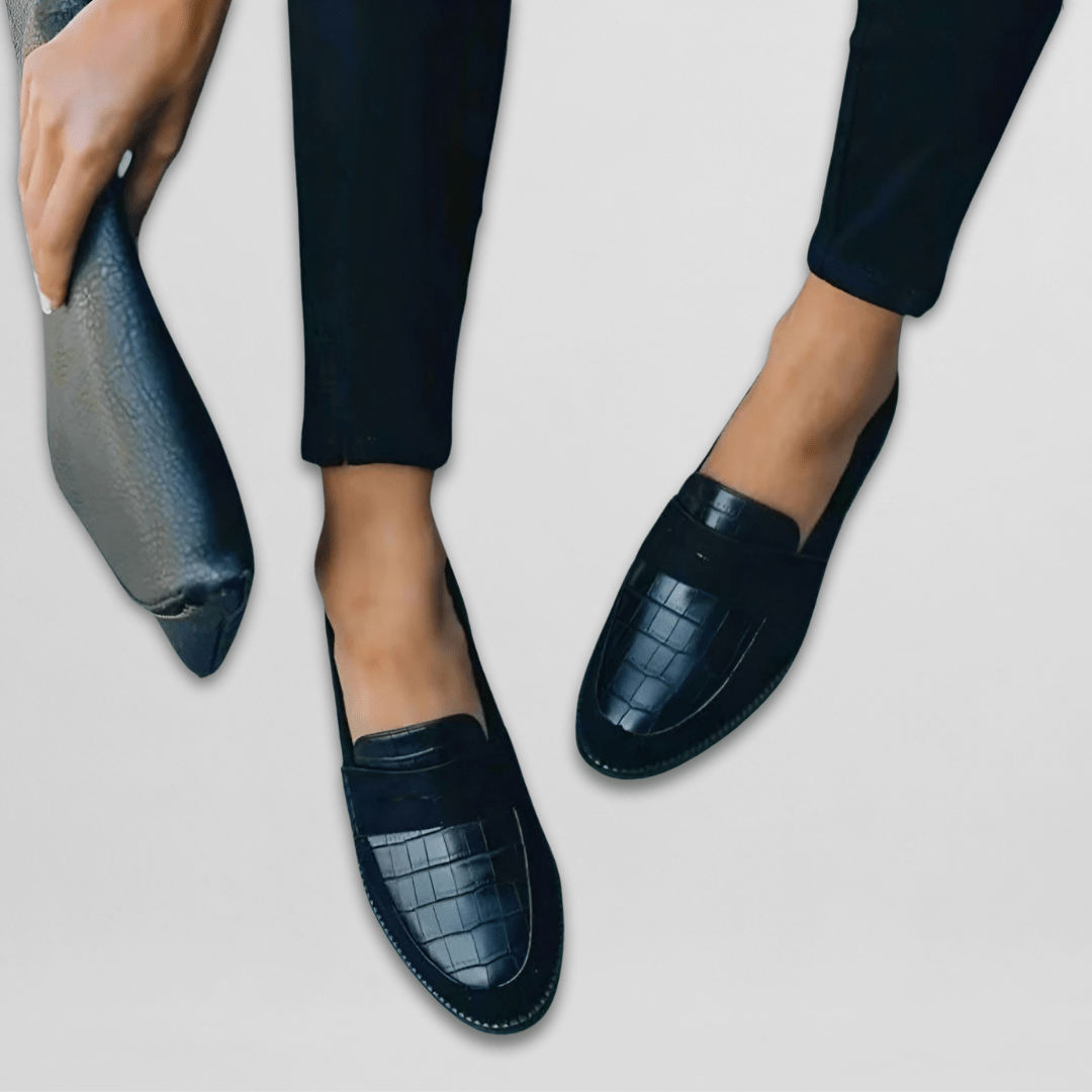 Mocasines negros colocados en diagonal, con detalle del relieve de efecto cocodrilo, la banda frontal y el perfil de tacón bajo; junto a ellos se muestra una suela.