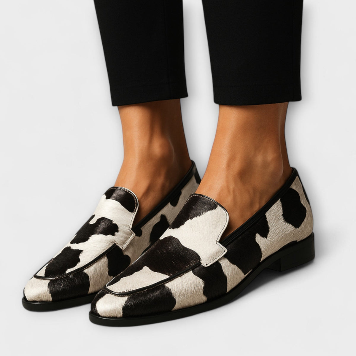 Mocasines de punta estilizada con estampado animal en blanco y negro, calzados con pantalones negros y vistos de frente en ángulo.