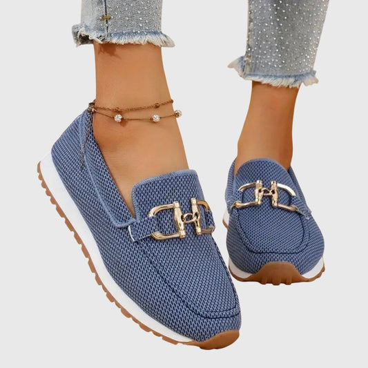 Mocasines deportivos azules con adorno metálico dorado, vistos en los pies desde una perspectiva frontal elevada.