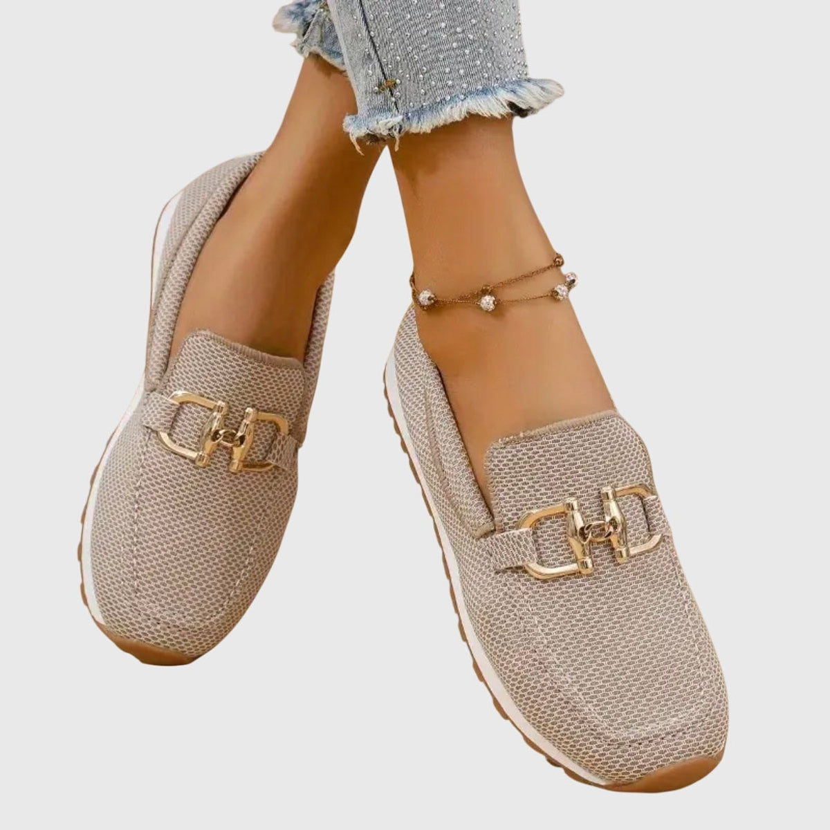 Mocasines deportivos beige con adorno metálico dorado, mostrados cruzados mientras se llevan puestos.