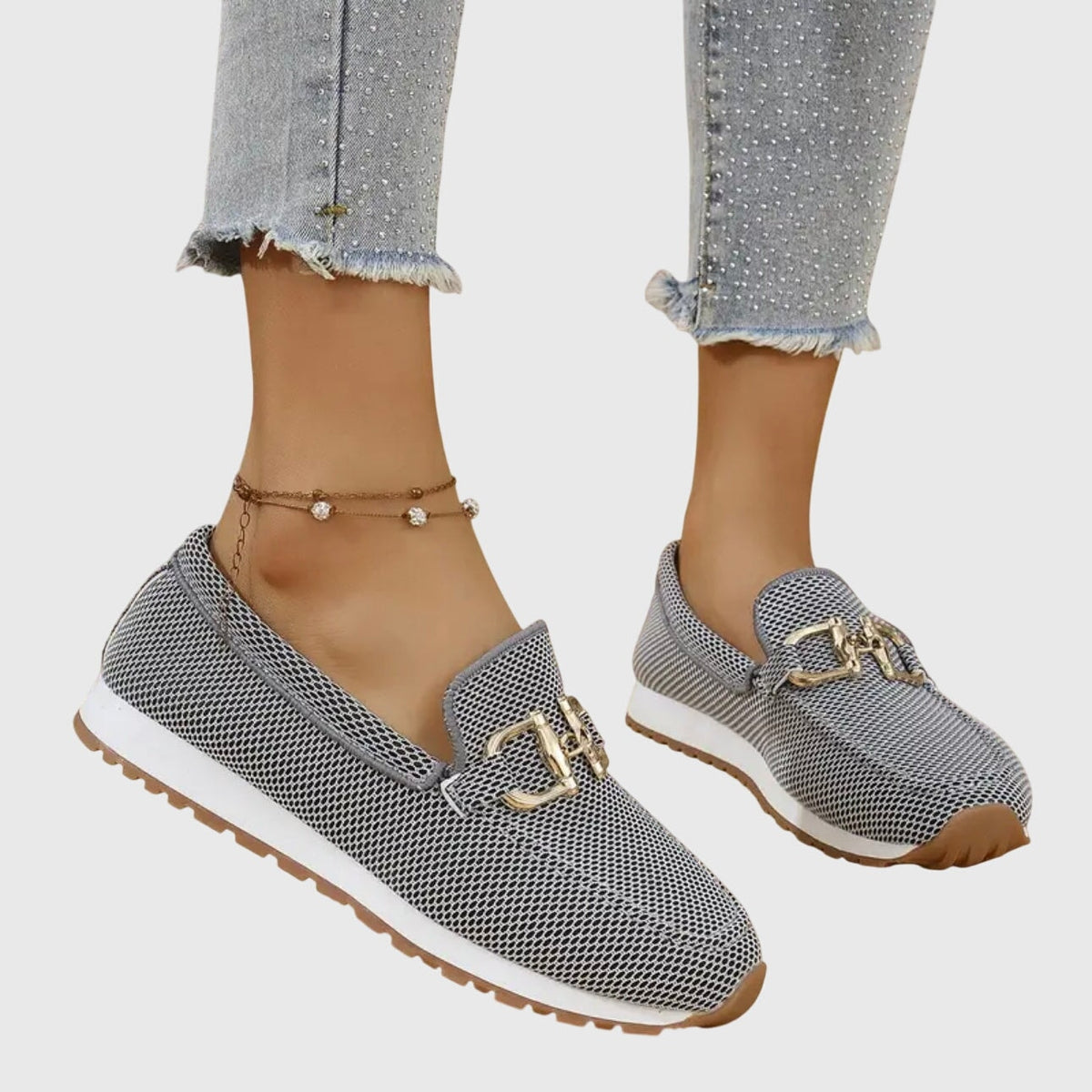 Mocasines deportivos grises con adorno metálico dorado y suela blanca, vistos en perspectiva lateral.