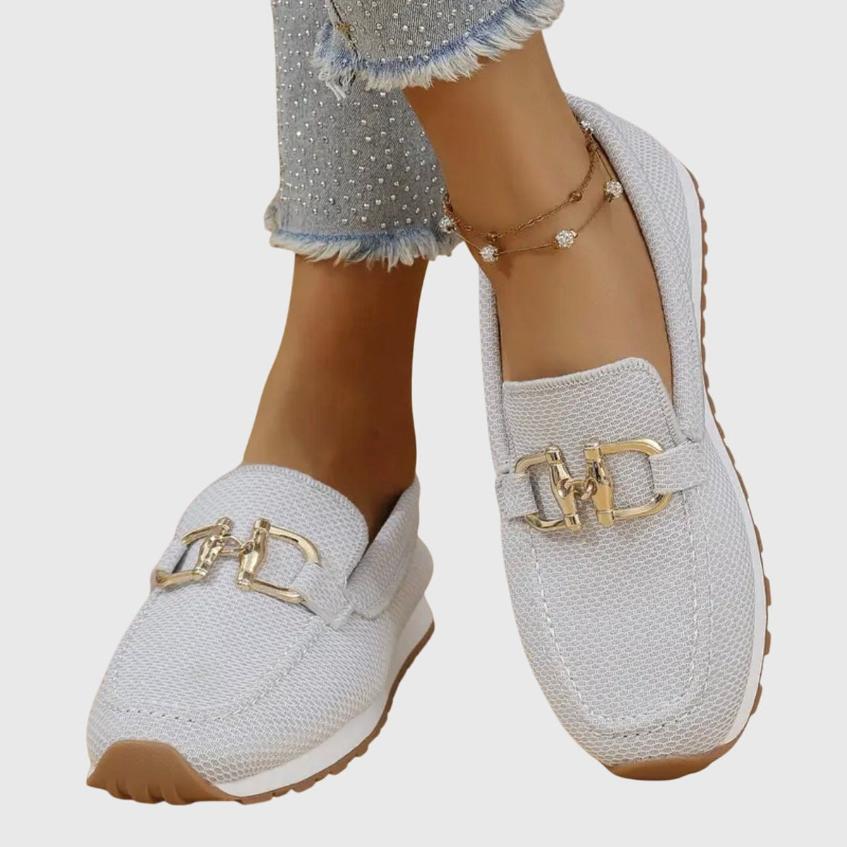 Mocasines deportivos blancos con adorno metálico dorado, mostrados de frente sobre los pies.