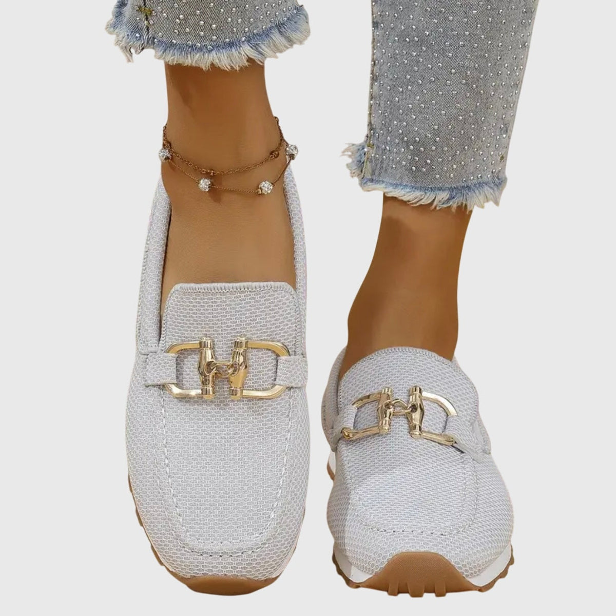 Mocasines deportivos blancos con adorno metálico dorado, vistos de frente en ambos pies.