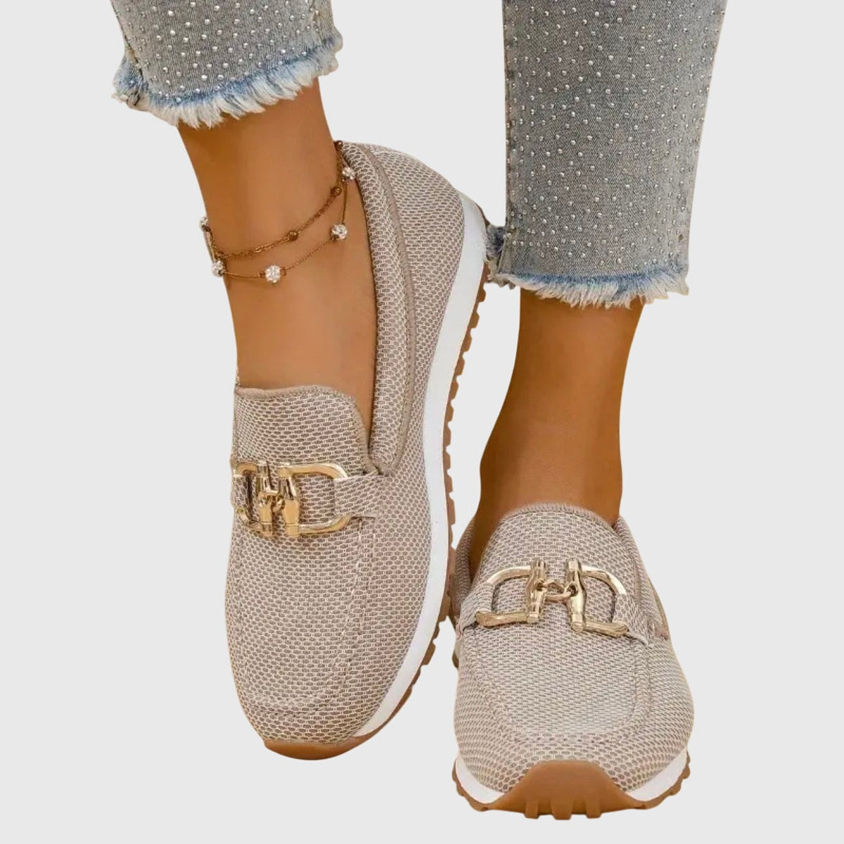 Mocasines deportivos beige con suela dentada marrón y adorno metálico, mostrados desde una vista frontal lateral.