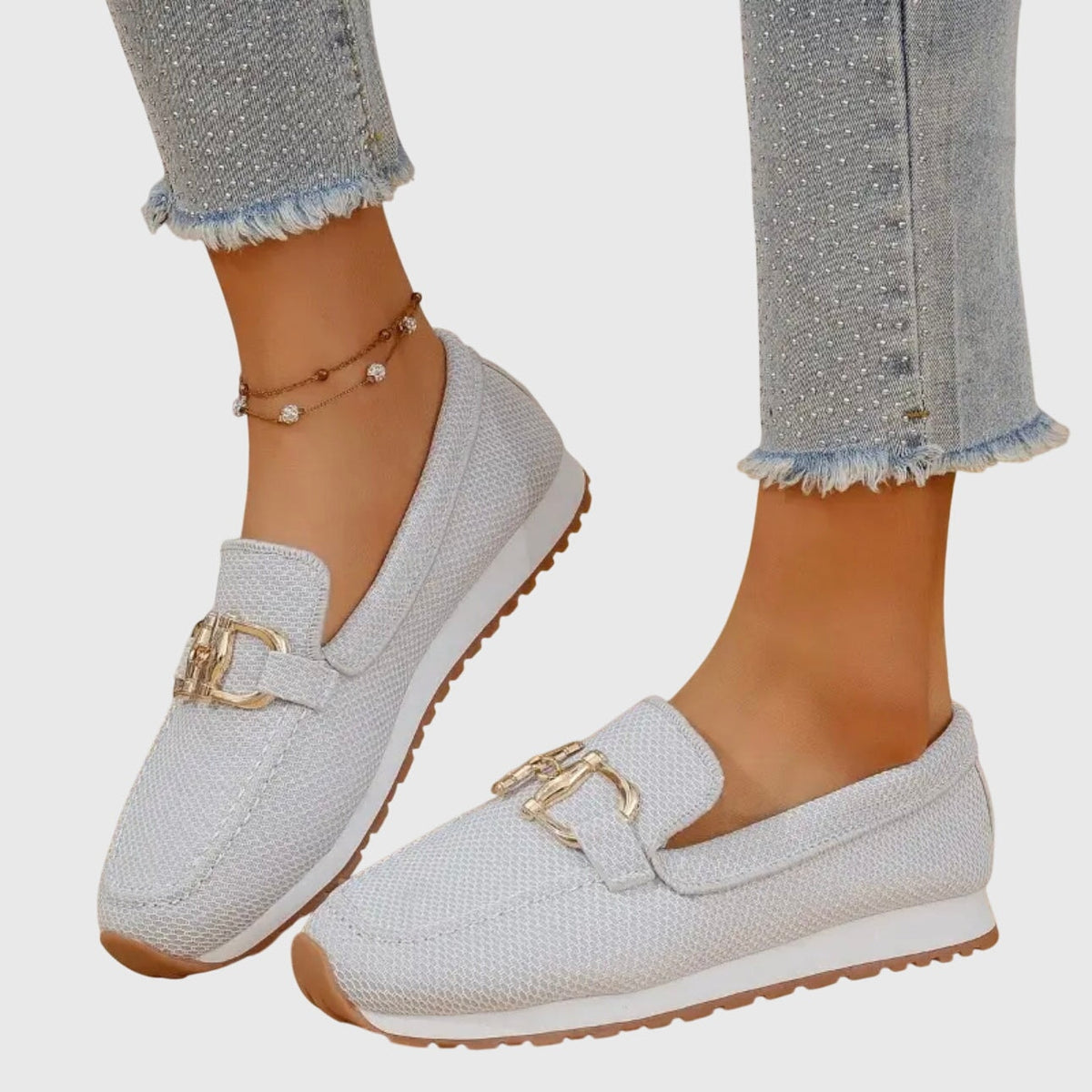 Mocasines deportivos blancos vistos de perfil en ambos pies, con suela blanca y detalle metálico.