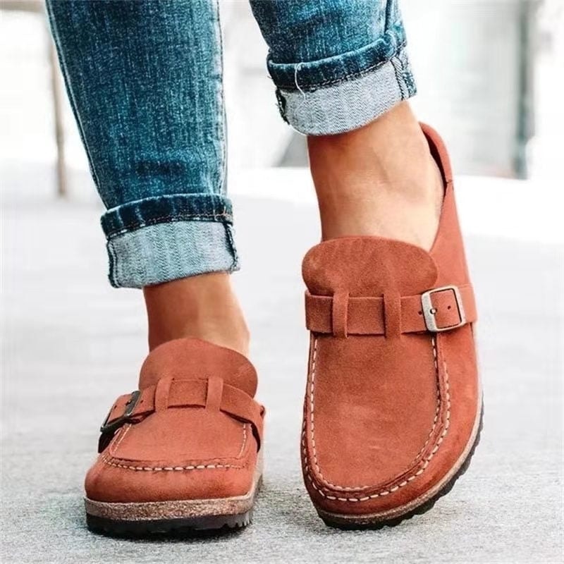 Mocasines planos en tono terracota, mostrados en los pies con vaqueros y hebilla ajustable en la tira frontal.