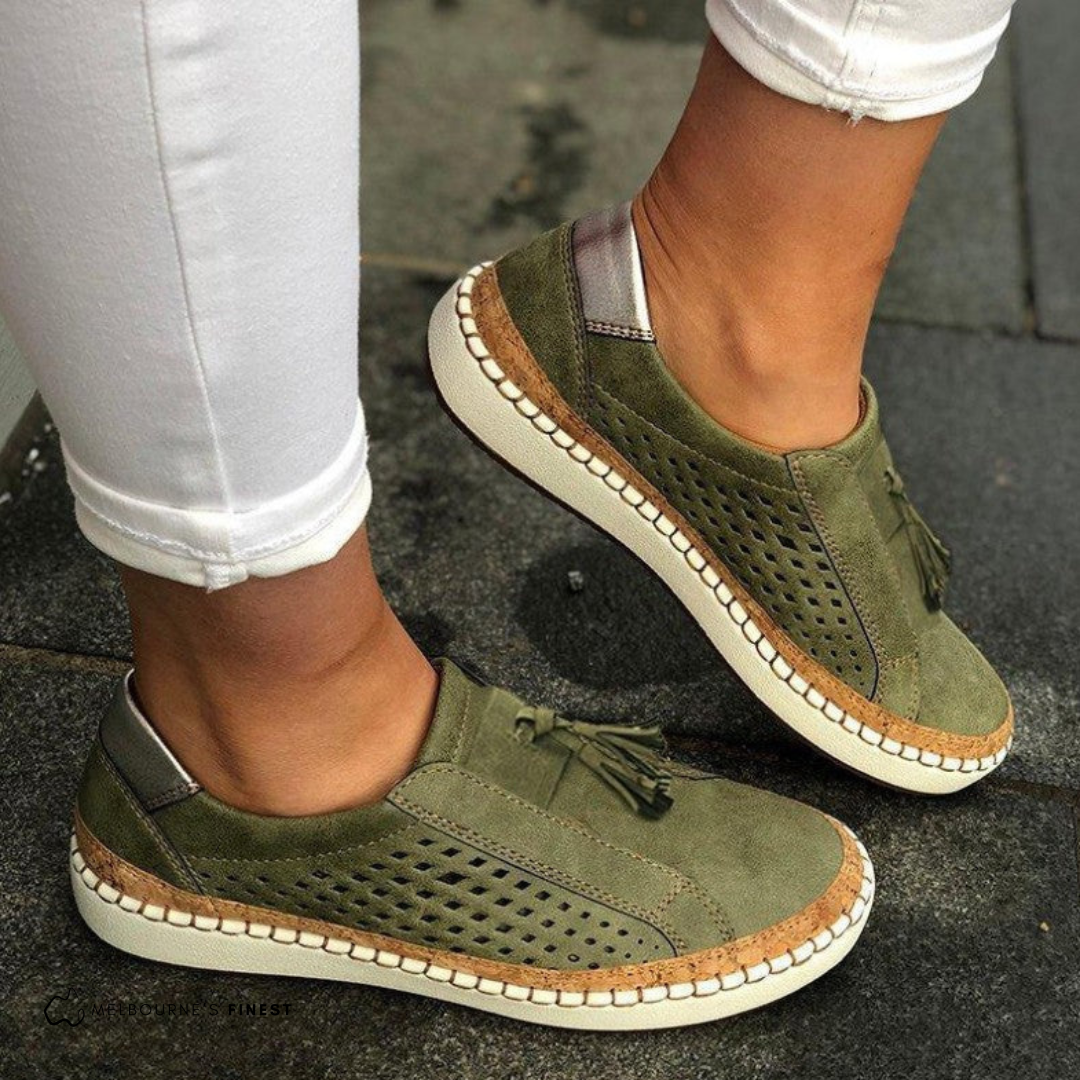 Mocasines planos verdes con laterales perforados, borlas decorativas y suela clara, vistos en los pies.