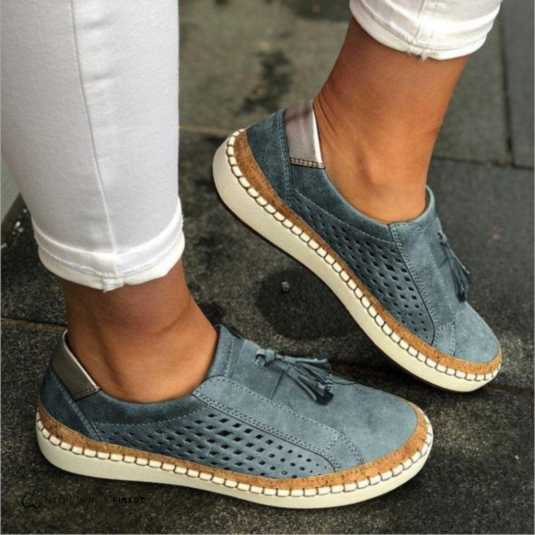 Mocasines planos azul grisáceo con diseño perforado, borlas en la parte superior y suela clara.