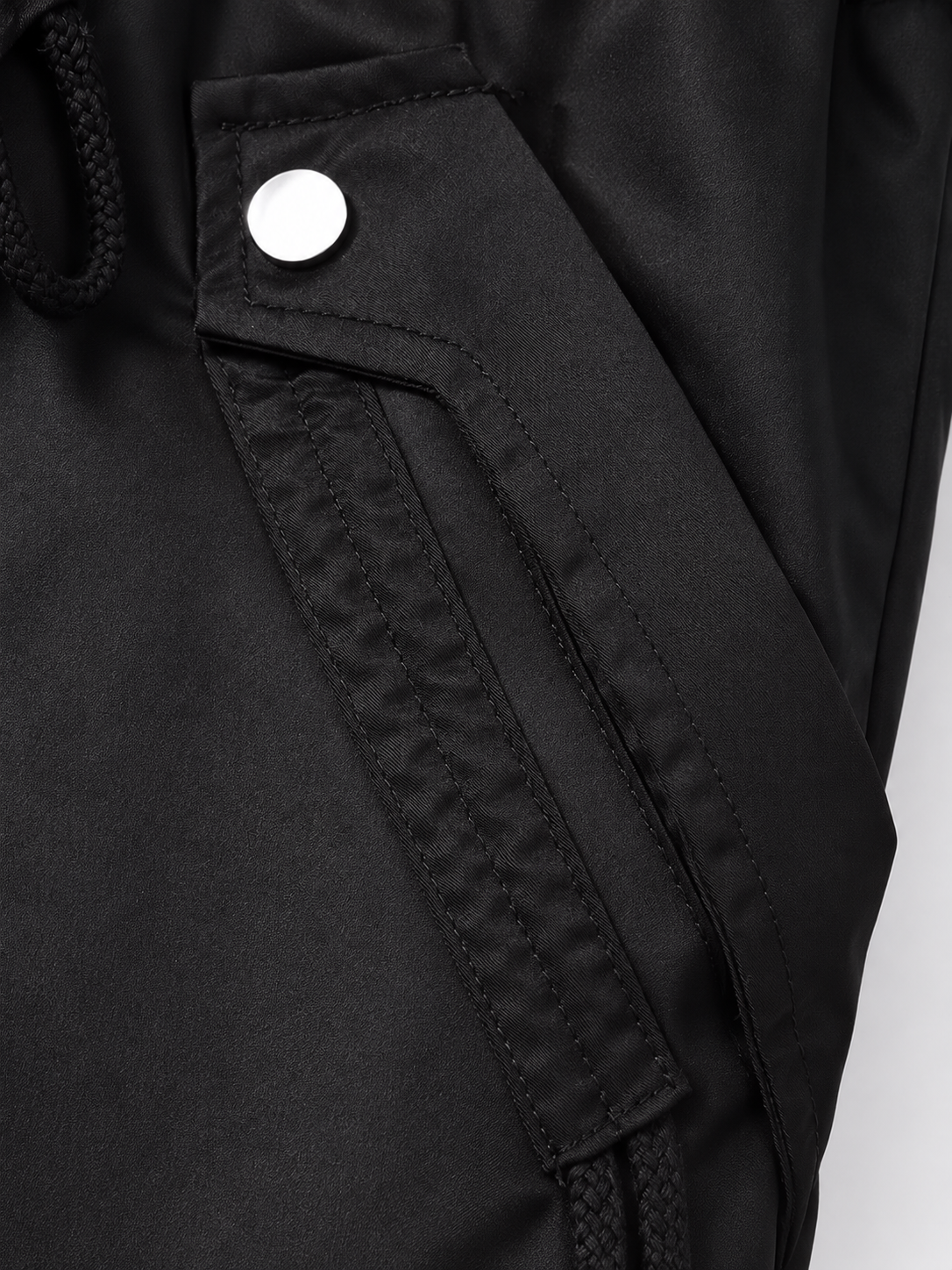 Detalle de un bolsillo lateral de la parka negra, con solapa, broche metálico y cordón de ajuste visible.