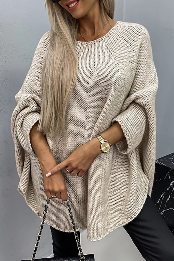 Poncho de punto holgado en tono beige, con cuello redondo, mangas largas remangadas y bajo asimétrico, combinado con pantalones negros.