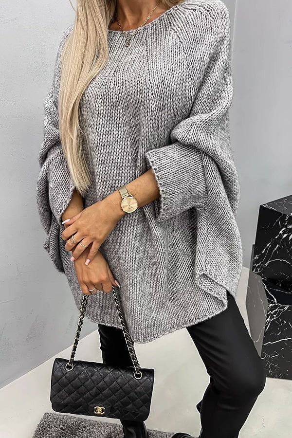 Poncho de punto holgado gris con cuello redondo y mangas amplias dobladas, mostrado con un bolso negro acolchado y pantalones oscuros.
