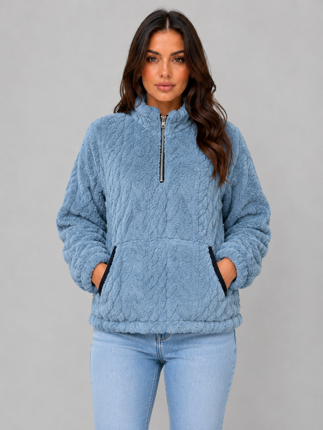 Sudadera azul de pelo con cuello alto, media cremallera y las manos dentro del bolsillo delantero.