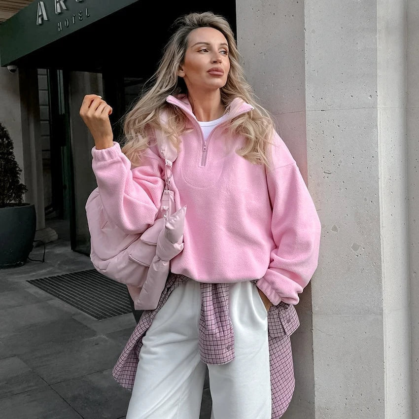 Mujer llevando la sudadera rosa de cuello alto y media cremallera con pantalones claros y un bolso, en un conjunto informal.