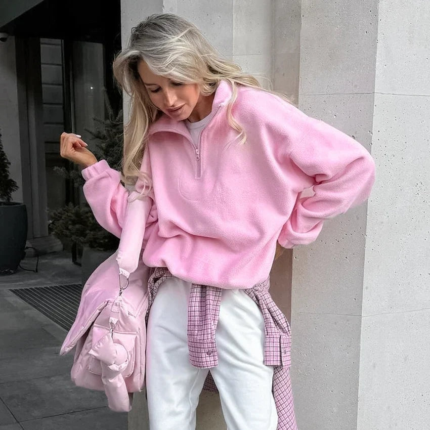 Mujer luciendo la sudadera rosa holgada con la media cremallera parcialmente abierta y el bajo recogido, en una pose casual.