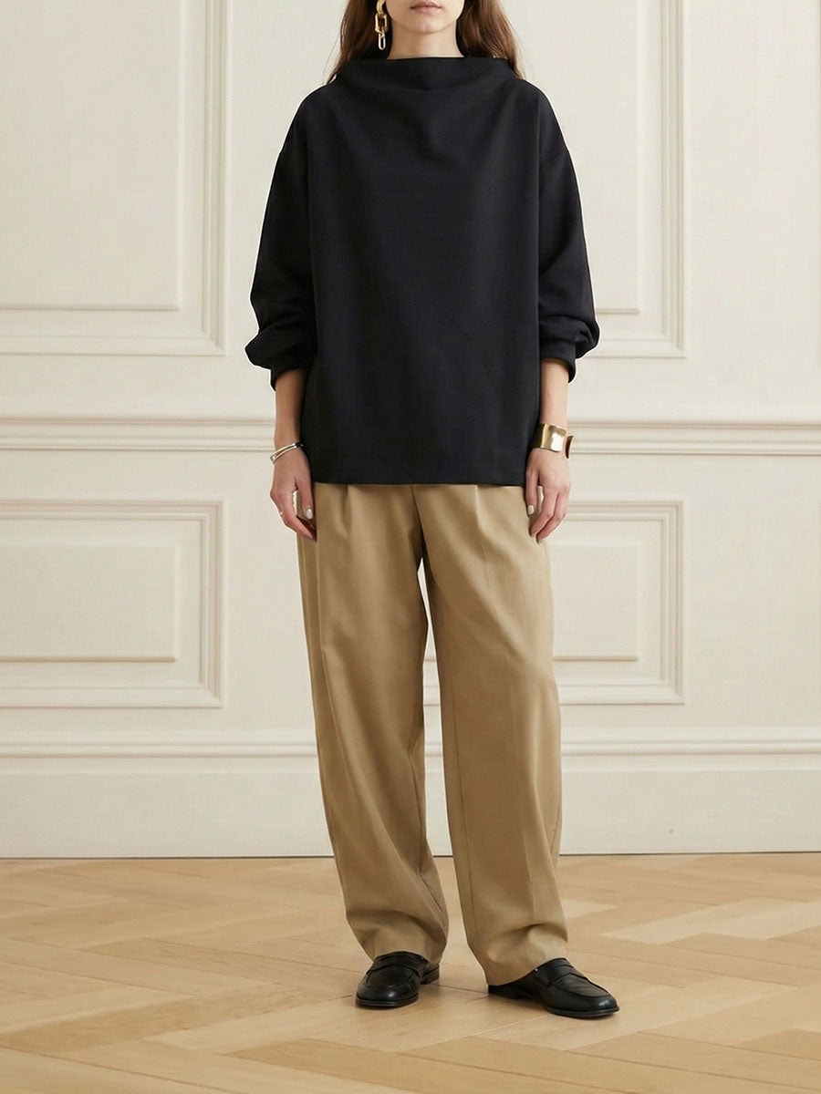 Vista de cuerpo entero de la sudadera negra de silueta relajada, cuello alto y mangas remangadas, combinada con pantalón beige.
