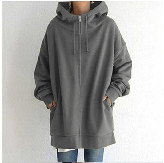 Sudadera oversize gris con capucha ajustable, cremallera frontal y bolsillos laterales, llevada con las manos dentro de los bolsillos.