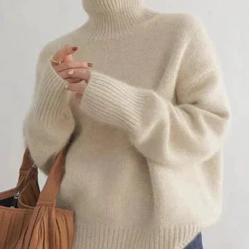 Suéter beige de cuello alto y corte holgado, con mangas largas y puños amplios, combinado con un bolso marrón.