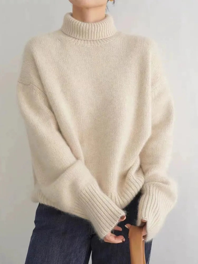 Suéter beige holgado de cuello alto y mangas largas, mostrado de frente con las manos juntas sobre unos vaqueros.