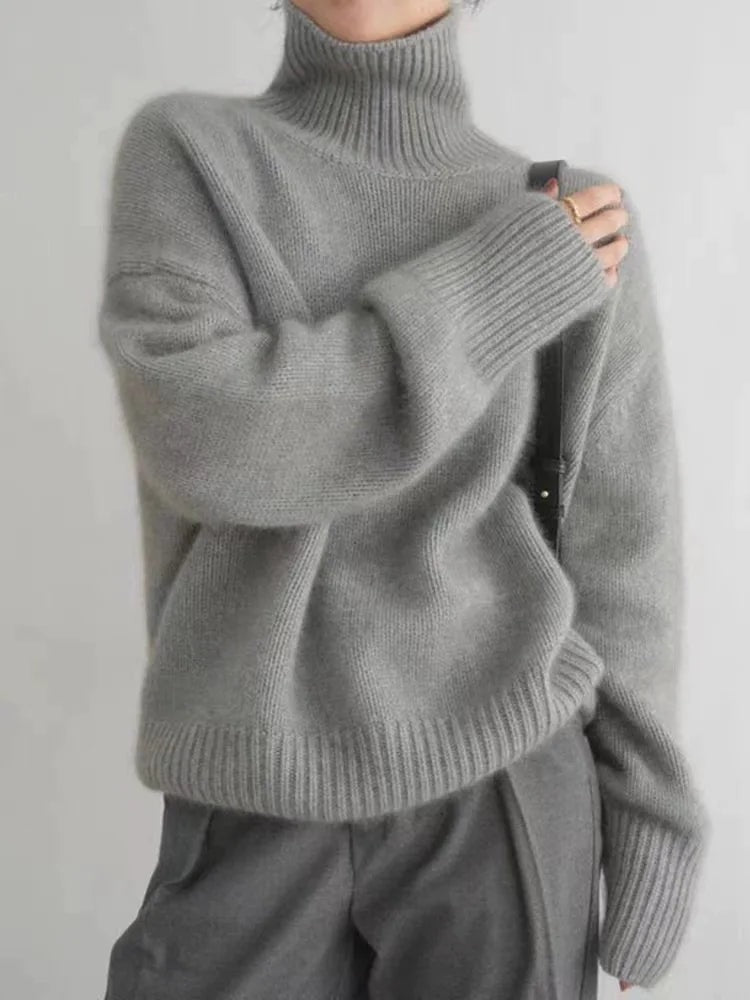 Suéter gris de cuello alto, hombros relajados y mangas largas, combinado con pantalones grises.