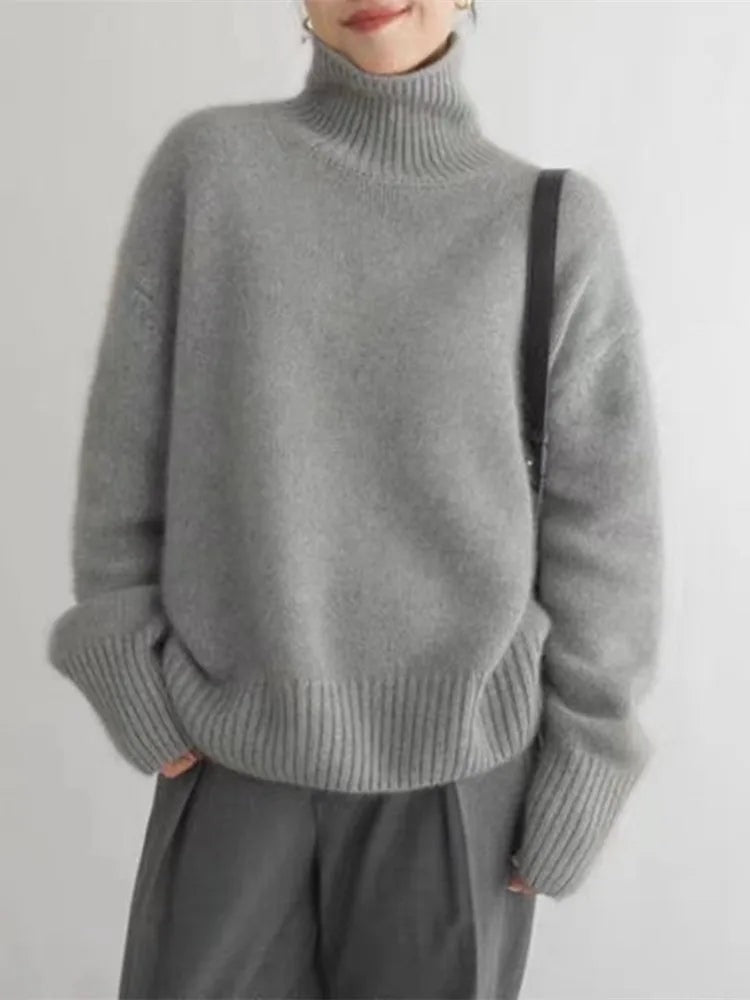 Vista frontal del suéter gris de cuello alto con silueta amplia y puños largos, combinado con pantalones de vestir.