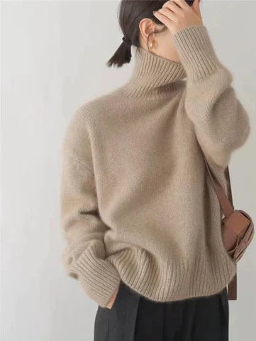 Suéter beige de cuello alto y corte holgado, visto de lado con un bolso pequeño al hombro y pantalón oscuro.