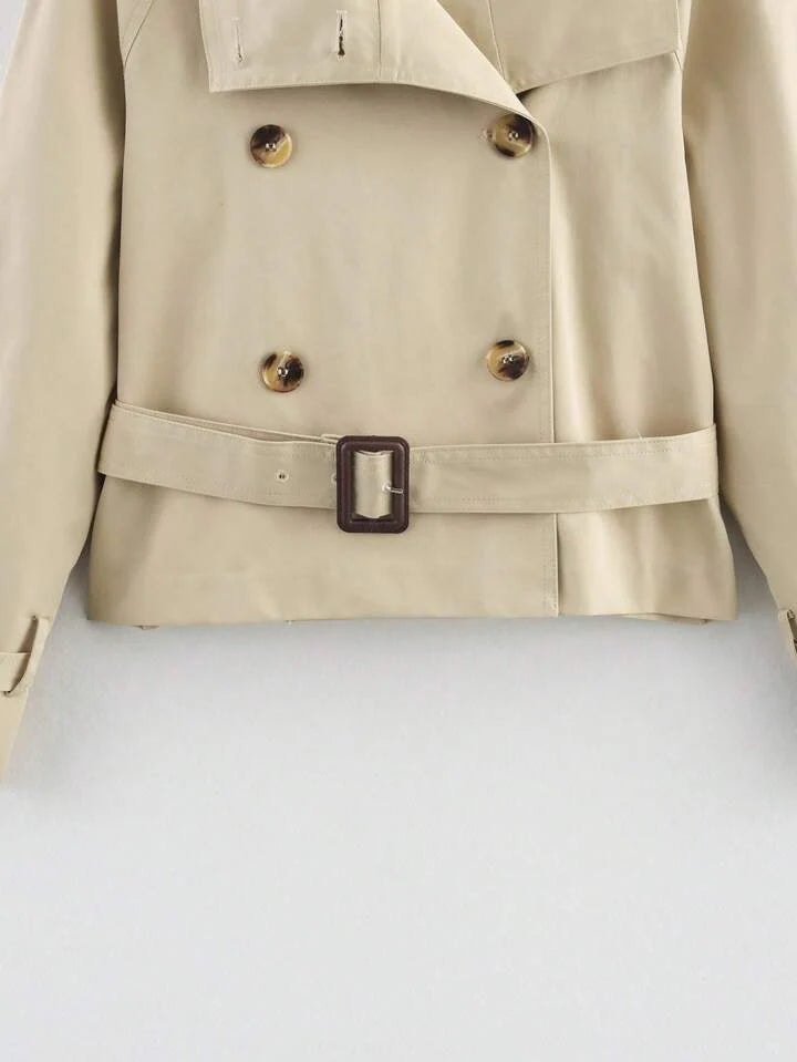Detalle frontal de la parte inferior del trench beige, mostrando los cuatro botones y el cinturón con hebilla rectangular.