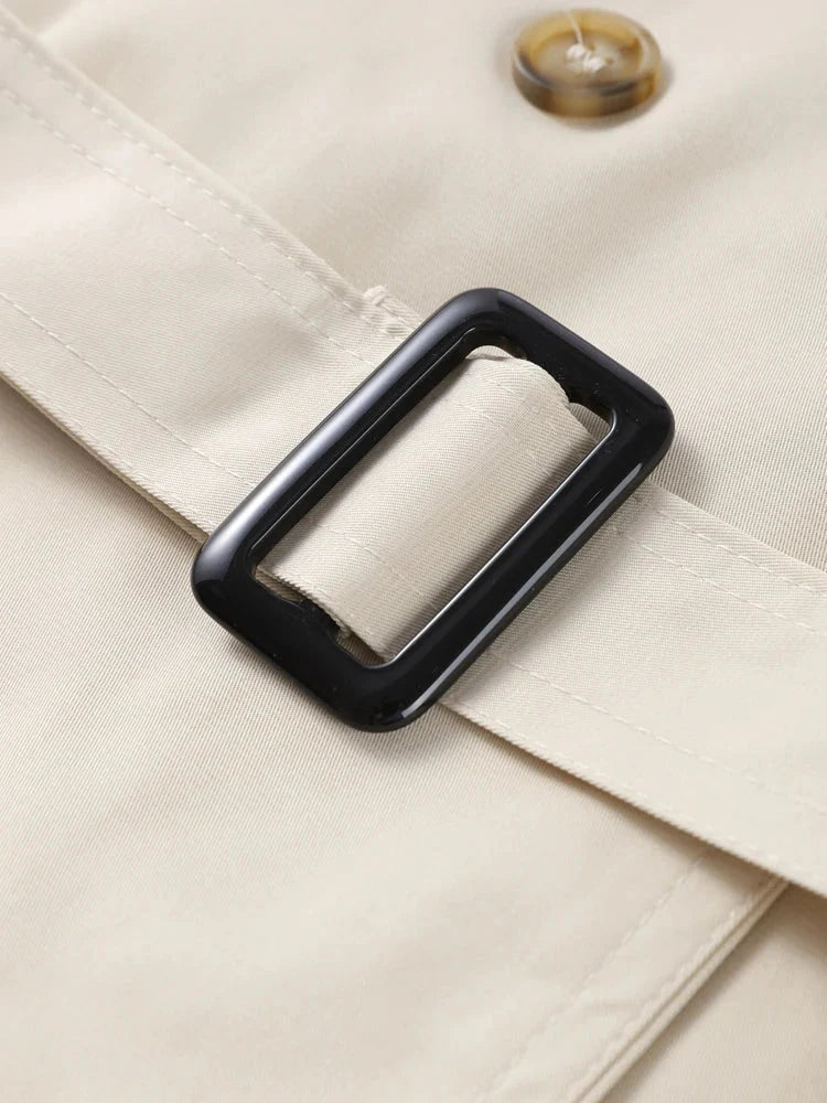 Primer plano de la hebilla rectangular oscura del cinturón sobre el trench beige, junto a uno de sus botones.