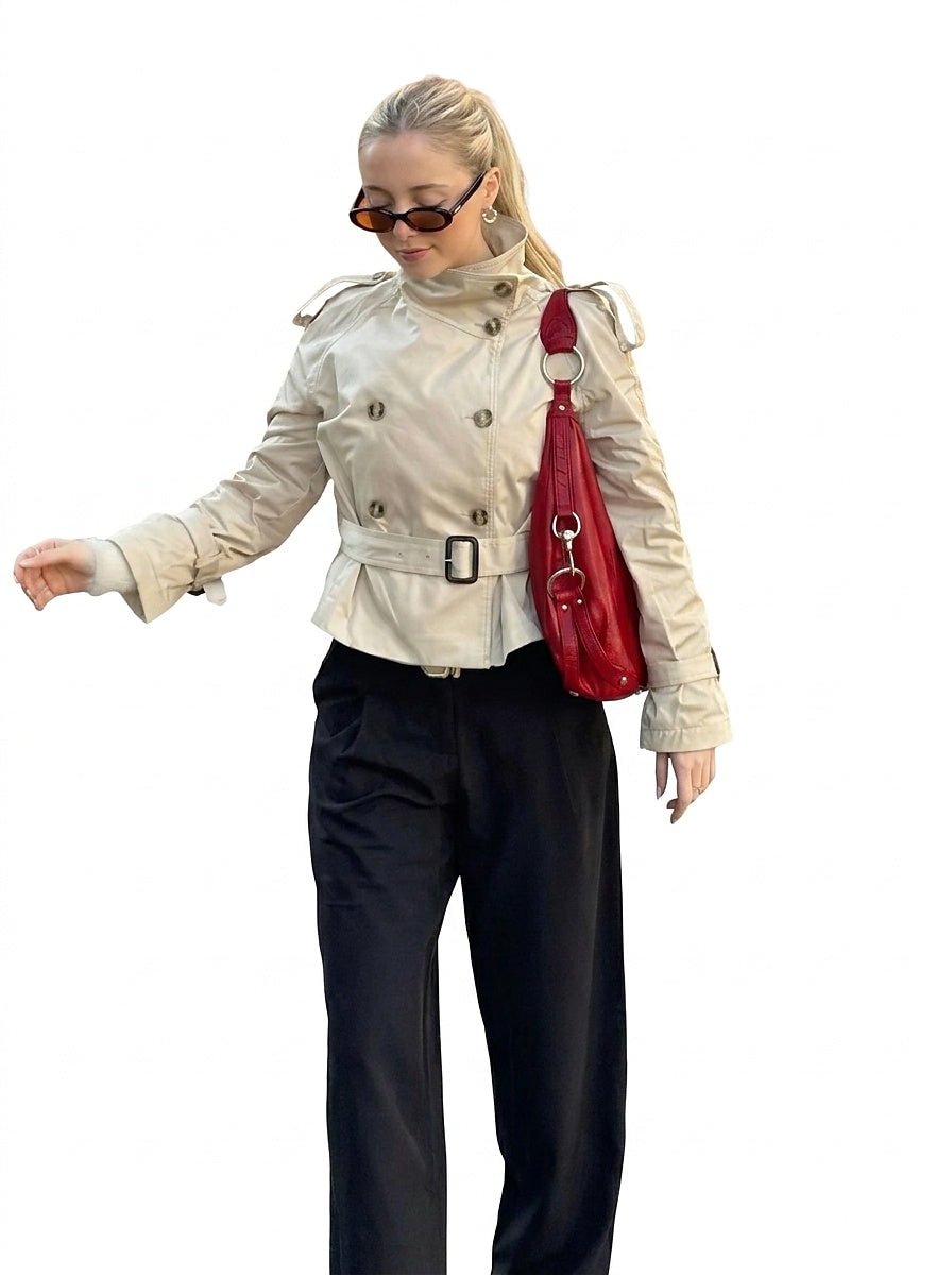 Modelo con trench corto beige abrochado, cinturón ceñido y cuello alto, combinado con pantalón oscuro y bolso rojo.
