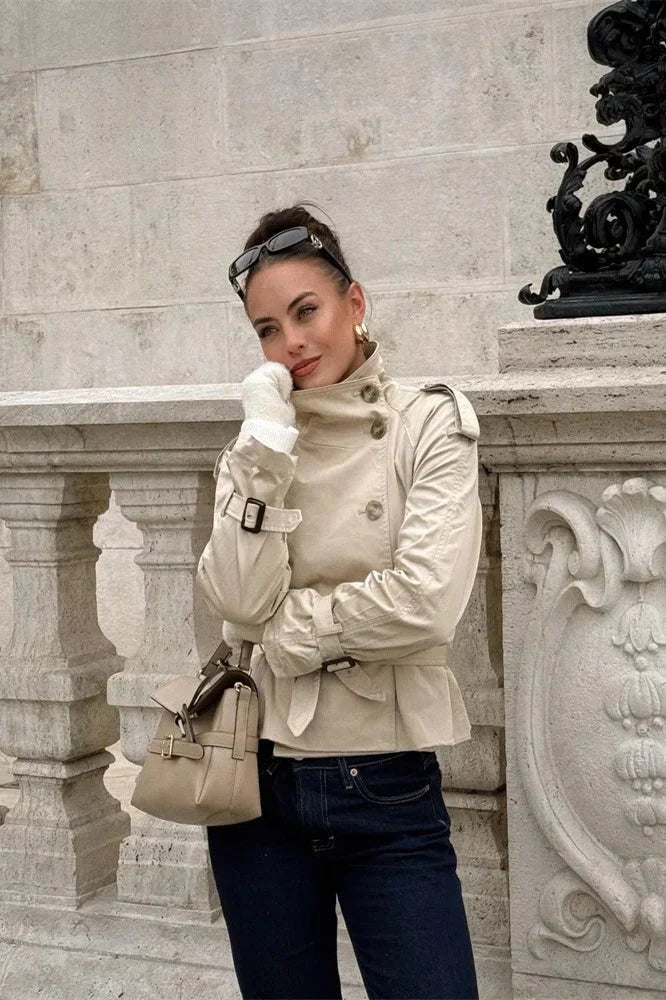 Trench corto beige llevado con el cinturón ajustado, mostrando el cuello alto, los detalles de los hombros y los puños abrochados.
