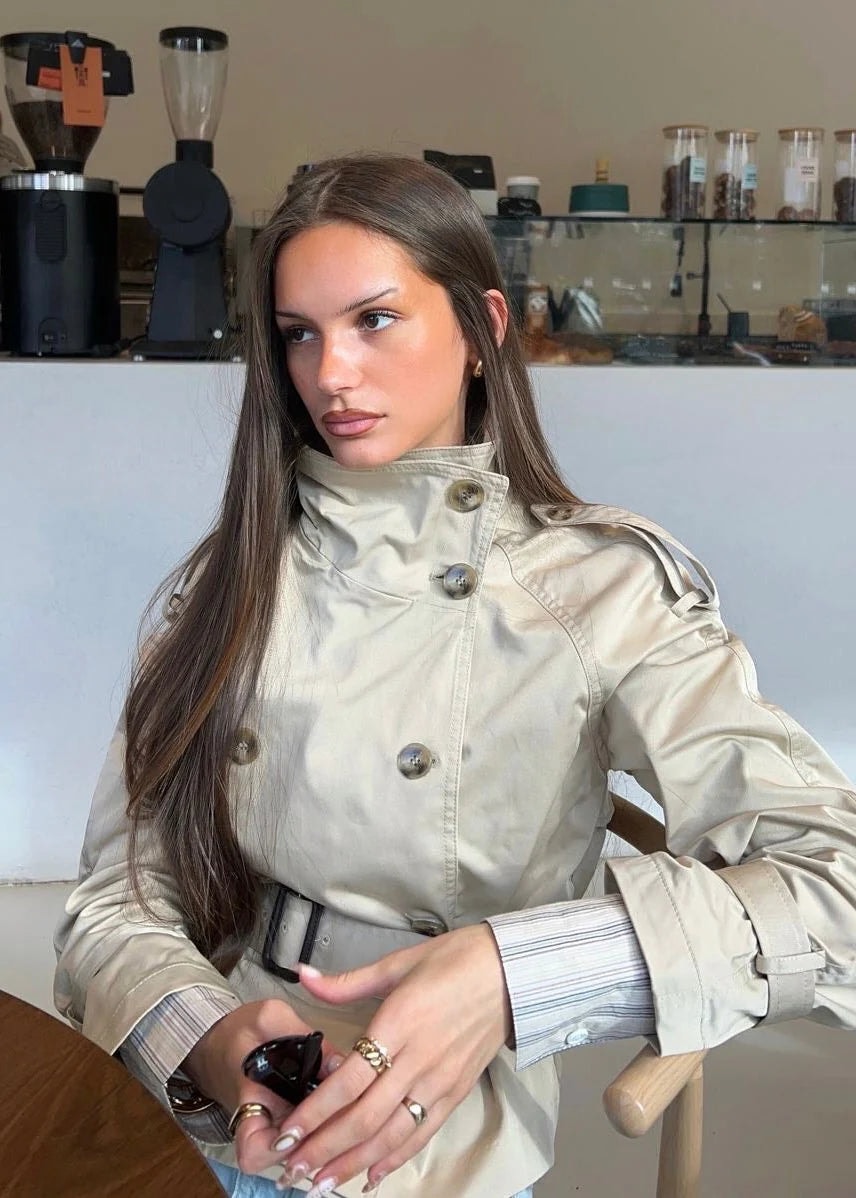 Detalle del trench beige llevado en interior, donde destacan el cuello alto abrochado, la doble botonadura y el cinturón a la cintura.