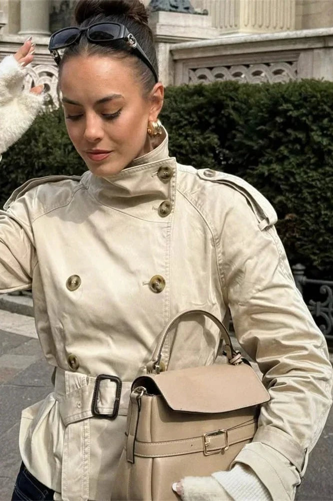 Modelo al aire libre con trench corto beige y bolso, mostrando los botones frontales, las hombreras con trabillas y el cinturón.