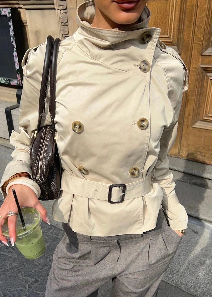 Acercamiento del trench corto beige cerrado, con cuatro botones visibles, cinturón de hebilla oscura y cuello alto.