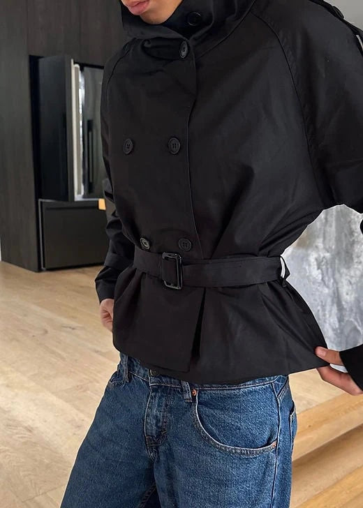 Trench corto negro llevado con vaqueros, visto de frente y con cinturón ajustado, doble botonadura y cuello alto.