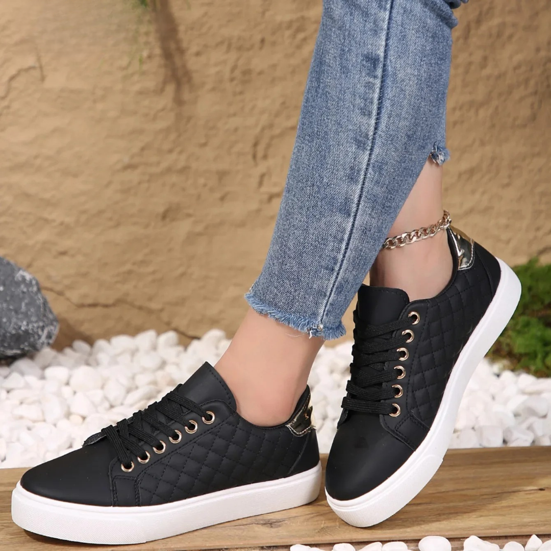 Zapatillas negras acolchadas con cordones, ojales dorados, suela blanca y detalle brillante en el talón, mostradas sobre una superficie de madera.