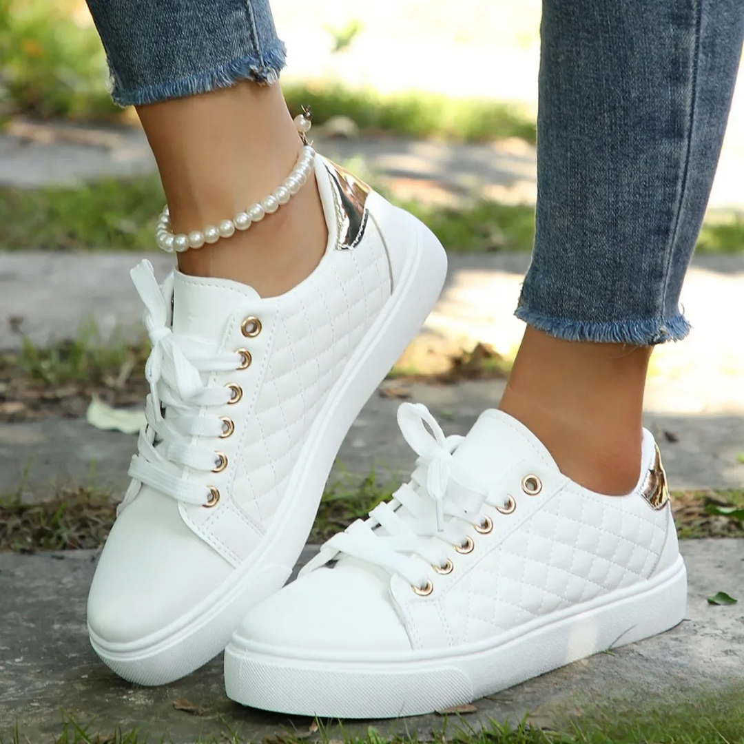 Zapatillas blancas de estilo casual con cordones, suela plana y acabados brillantes en el talón, combinadas con pantalones vaqueros de bajo desflecado.