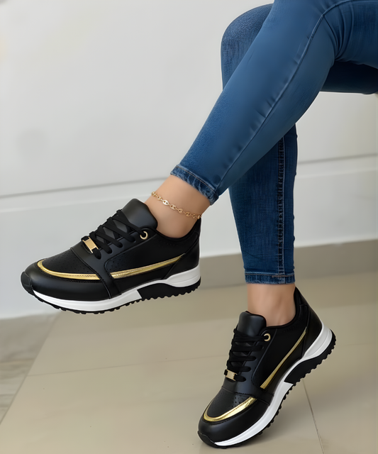 Zapatillas deportivas negras con detalles metalizados dorados, cordones negros y suela marcada blanca y negra, llevadas con vaqueros.