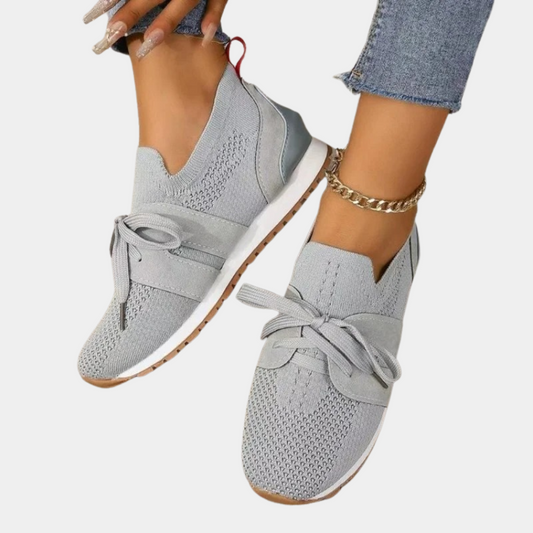 Zapatillas ligeras grises con empeine texturizado, cordones y suela clara, mostradas en los pies junto a unos vaqueros.
