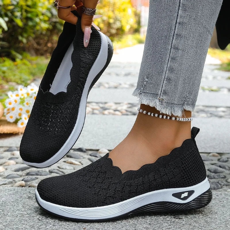 Zapatillas slip-on negras con borde ondulado y suelas blancas acolchadas, mostradas en ambos pies.