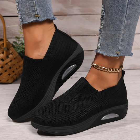 Zapatillas slip-on negras con suela gruesa y detalles transparentes en el lateral, puestas con vaqueros.