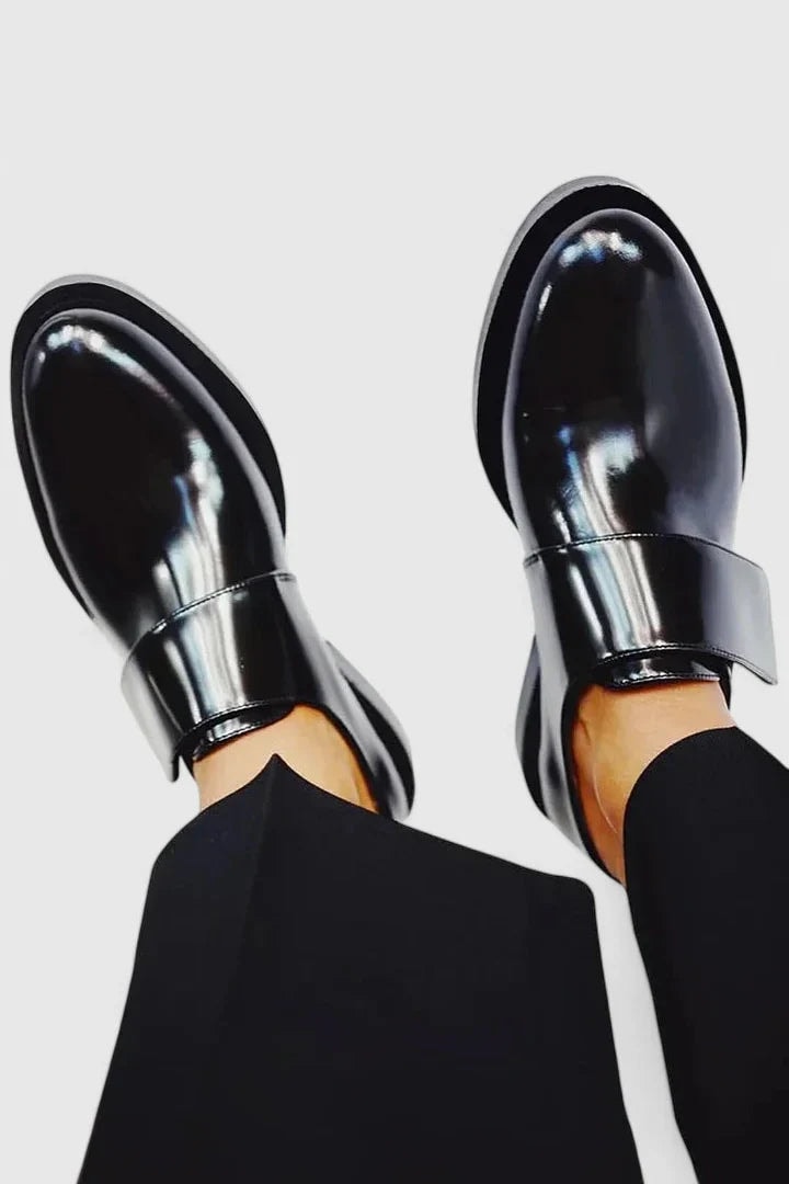 Vista superior de los zapatos planos negros brillantes mostrando las correas anchas y el perfil de los laterales mientras se llevan con pantalón oscuro.