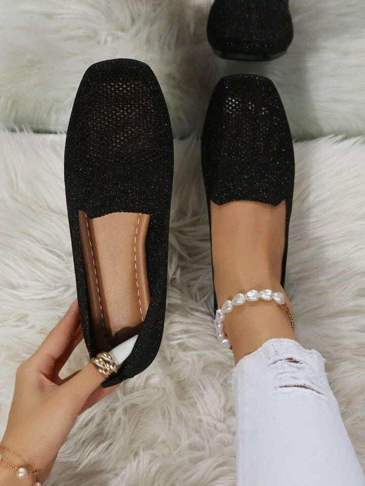 Zapatos planos negros de tejido calado, vistos desde arriba junto a un pie con tobillera de perlas.