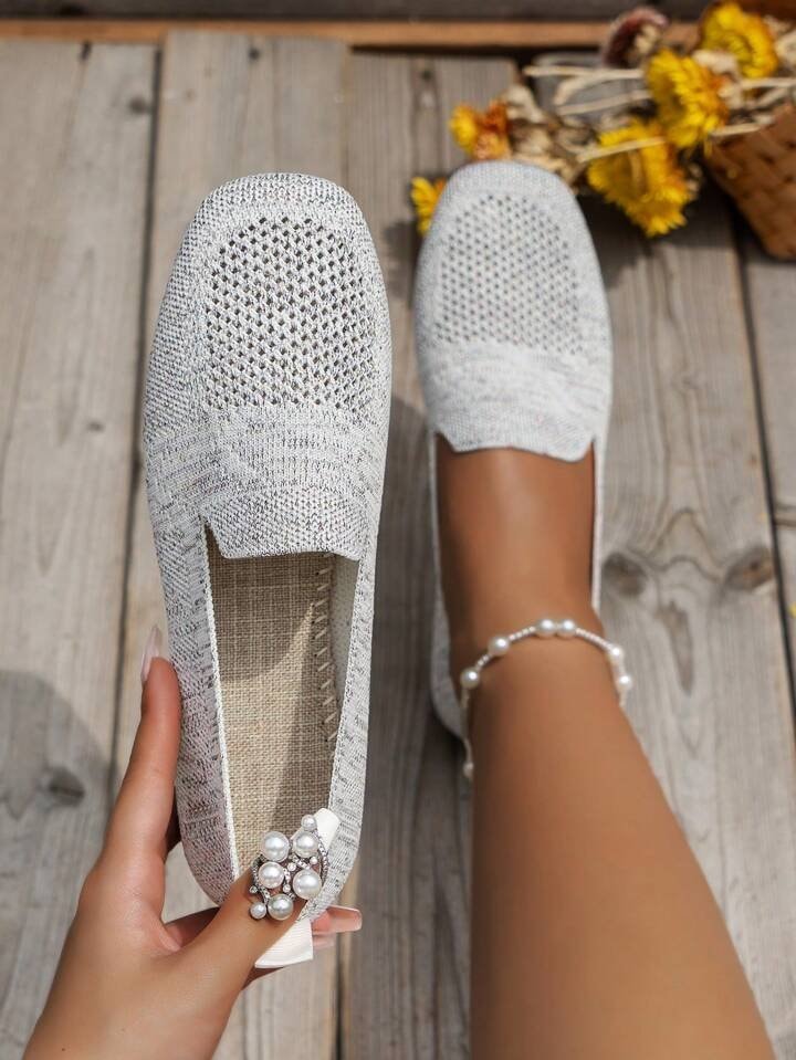 Zapatos planos blancos de diseño calado sobre tablas de madera, con detalle decorativo de perlas en uno de ellos.