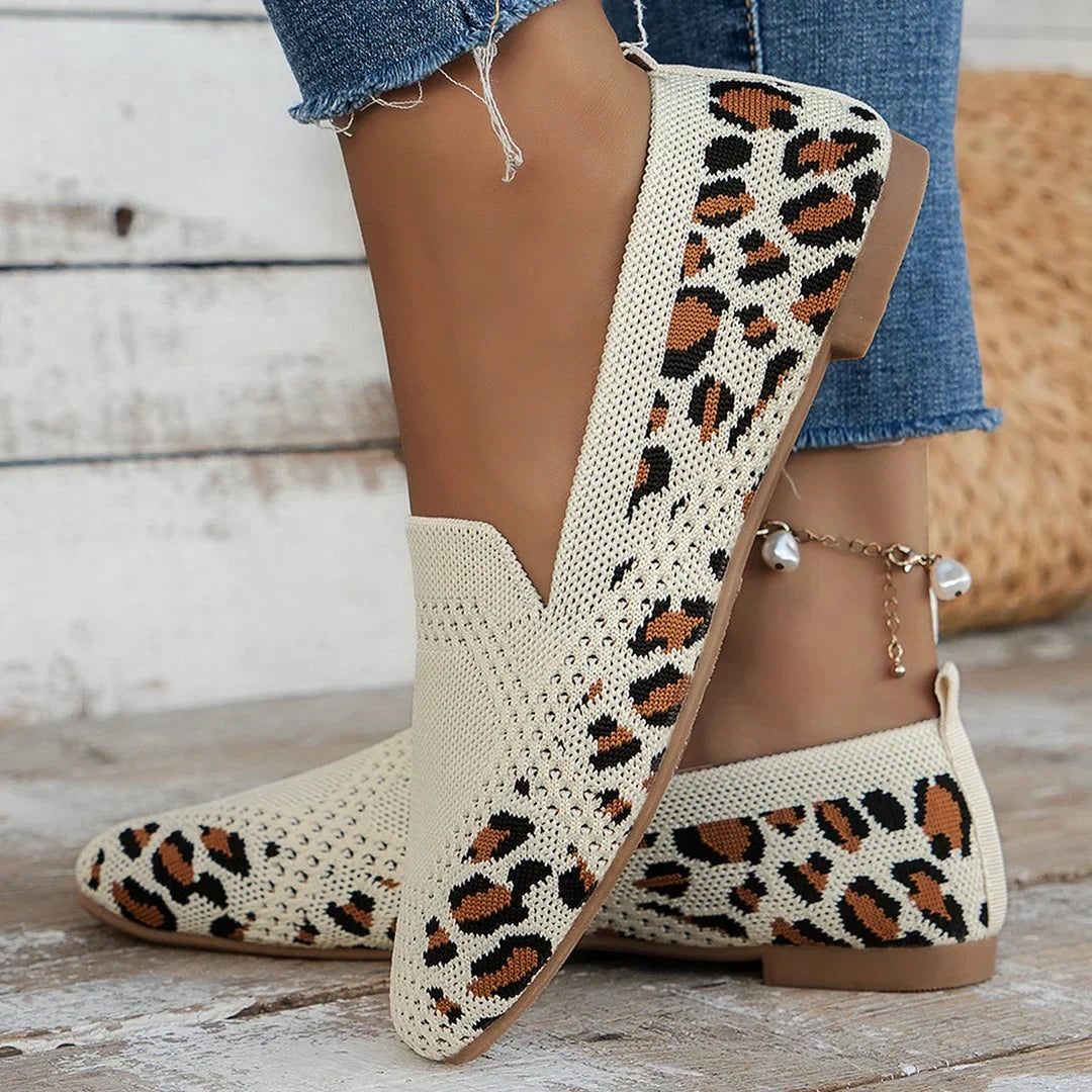 Zapatos planos con estampado animal en tonos crema, negro y naranja, vistos de lado sobre una superficie de madera.